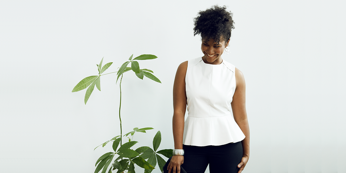  A smiling woman wearing a white sleeveless blouse and black pants stands next to a potted plant.